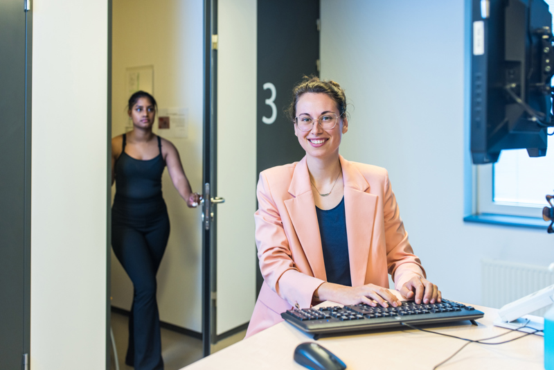 Een jeugdarts kijkt in de camera met haar vingers op het toetsenbord, terwijl een patiënt de kamer binnenloopt